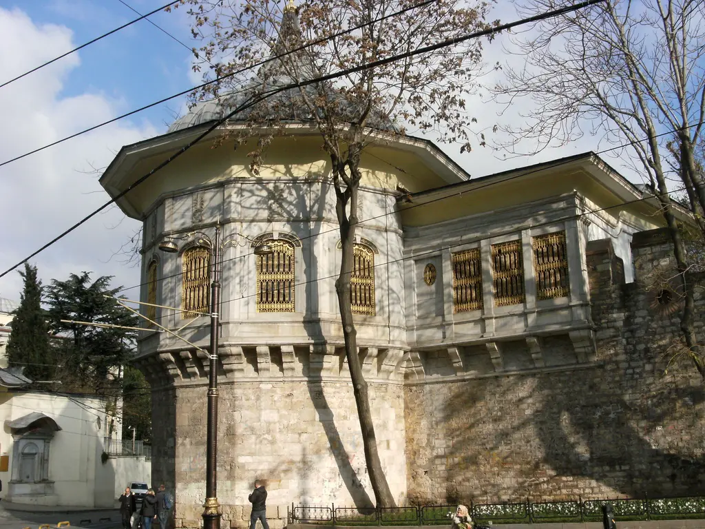 Alay Köşkü'nün sur köşesi; taş burcun üstünde mermer salon, altın sarısı pencere şebekeleri, yandaki kanat ve ağaç gölgeleri.