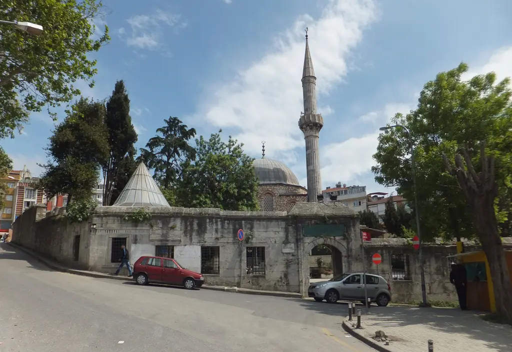 Çinili Camii'nin avlu duvarı; sokak kenarında taş duvar, arkasında şadırvanın konik çatısı, kubbe ve minare.