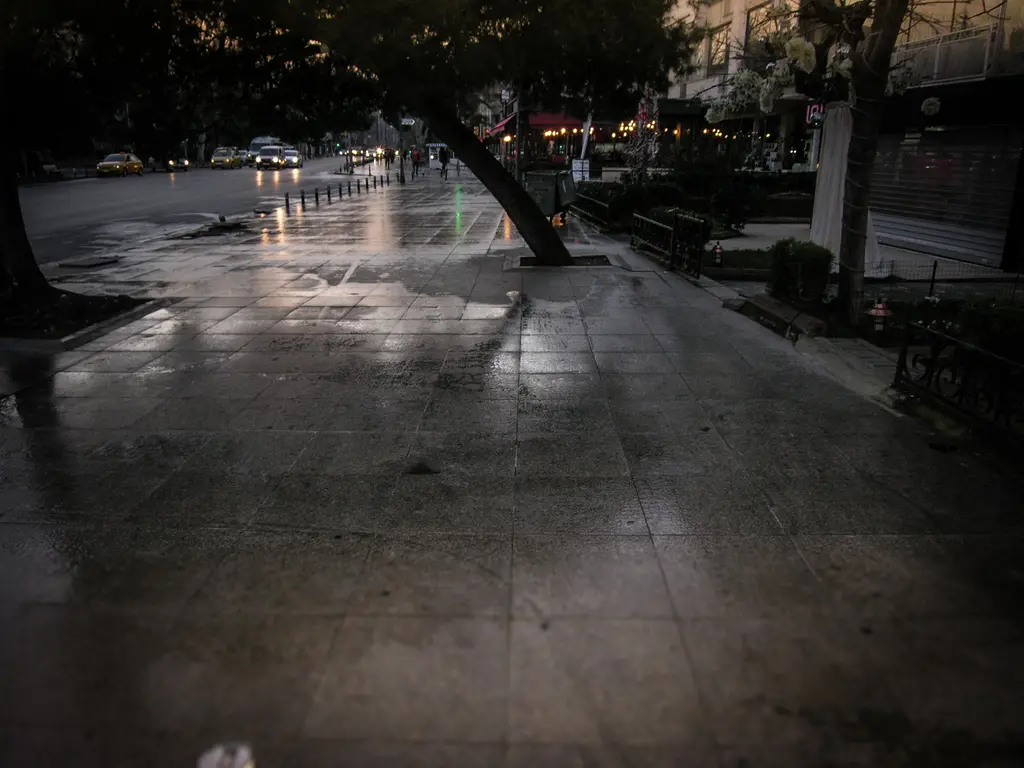 Yağmur sonrası Bağdat Caddesi'nde ıslak kaldırım; eğik bir ağaç, banklar, cadde lambaları ve arabalar.