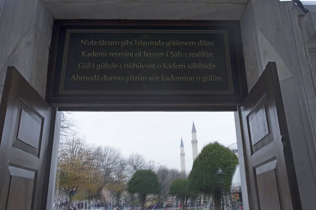 Sultan Ahmed Türbesi'nde çıkış kapısının üstü; siyah zemine yazılmış dört satırlık şiir levhası, açık kanatlar ve dışarıda meydandaki minareler.