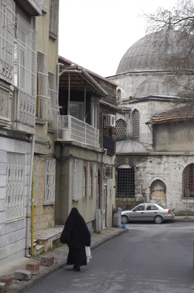 Haseki Sultan Külliyesi'ne çıkan dar sokak; iki yanda cumbalı eski evler, sonunda kurşun kubbeli taş yapı ve yürüyen bir kadın.