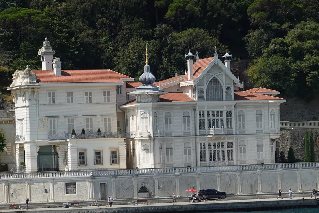 Huber Köşkü; beyaz ahşap cephe, kiremit çatılar, sivri kuleler, süslü pencereler ve arkada yeşil koru.