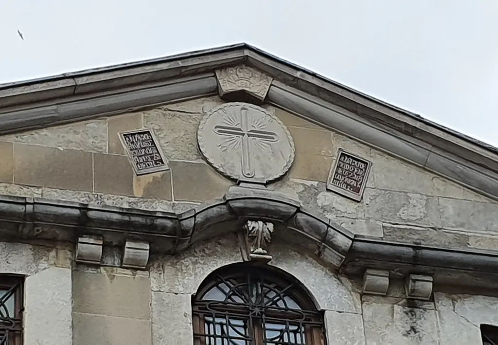 Aya Yorgi Kilisesi'nin cephe alınlığı alttan; ortada ışınlı haç işlenmiş yuvarlak taş madalyon, iki yanda Yunanca kitabe levhaları ve altta kemerli pencere.