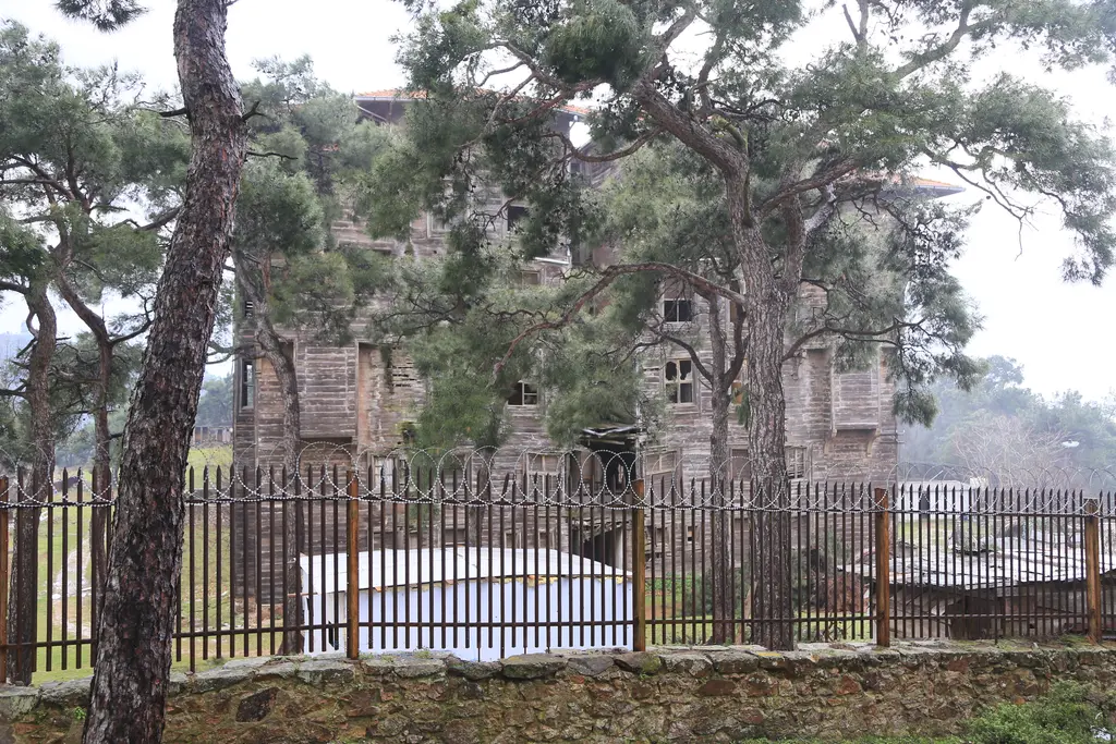Büyükada Rum Yetimhanesi; çam ağaçlarının ardında ahşap cephe, önde demir parmaklıklı çit.