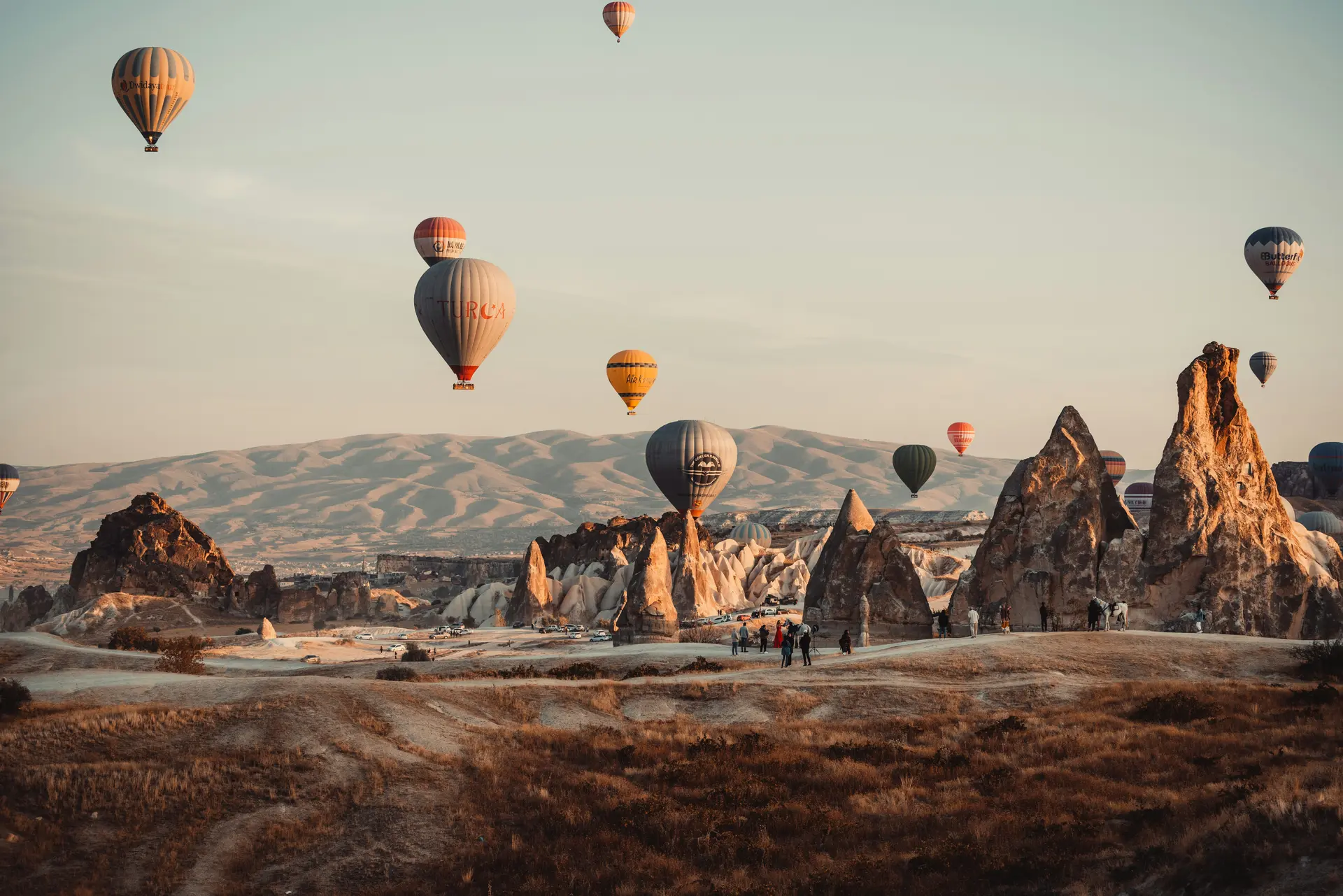 Kapadokya'da (Nevşehir) gün doğumunda peri bacaları vadisinin üzerinde süzülen onlarca rengarenk sıcak hava balonu; altın rengi ışıkta uzanan tüf kayalar ve arkadaki dağlar.