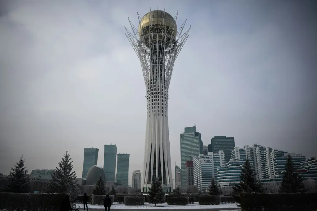 Astana'da kışın Bayterek Kulesi; beyaz çelik dalların taşıdığı altın küre, arkasındaki cam gökdelenler, karla kaplı zemin ve gri gökyüzü.