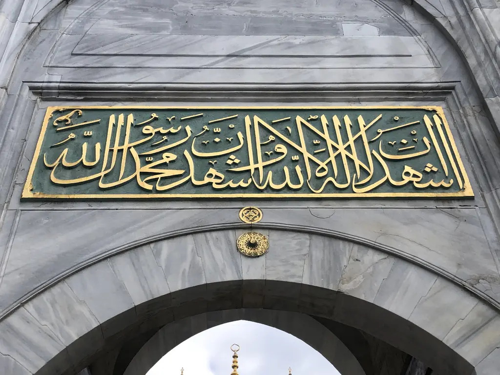 Yeni Valide Camii'nde yazı levhası; yeşil zemin üzerine altın harflerle Arapça yazı, altında mermer kemer.