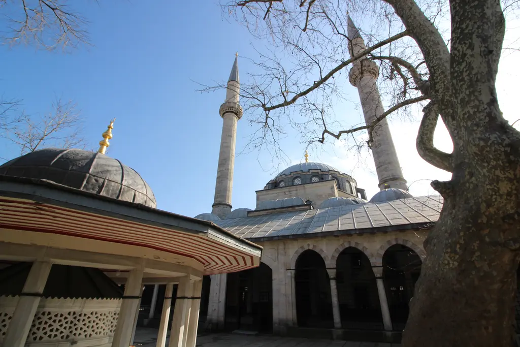 Atik Valide Camii'nin avlusu; kubbeli şadırvan, revaklar, arkada caminin kubbesi ve iki minaresi.