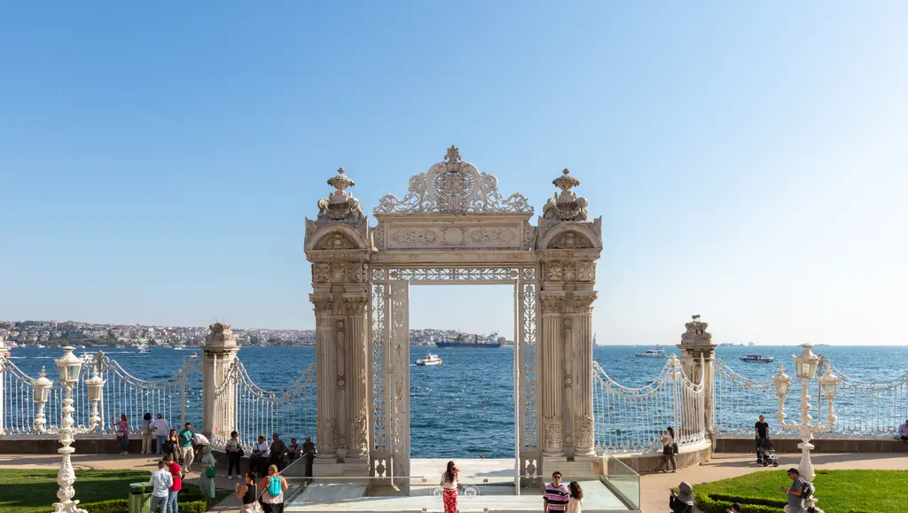 Dolmabahçe Sarayı'nın deniz kapısı; kıyıda süslü taş kemer, demir parmaklıklar, arkada Boğaz'ın suyu ve karşı kıyı.