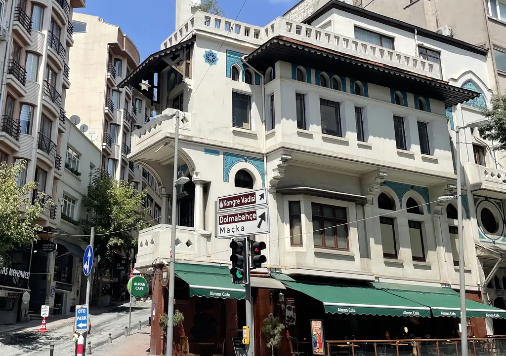 Nişantaşı'nda Vedat Tek Evi; çinili cumbalar, geniş saçaklı beyaz köşe cephesi, zemin katta yeşil tenteli kafe ve yön levhaları.
