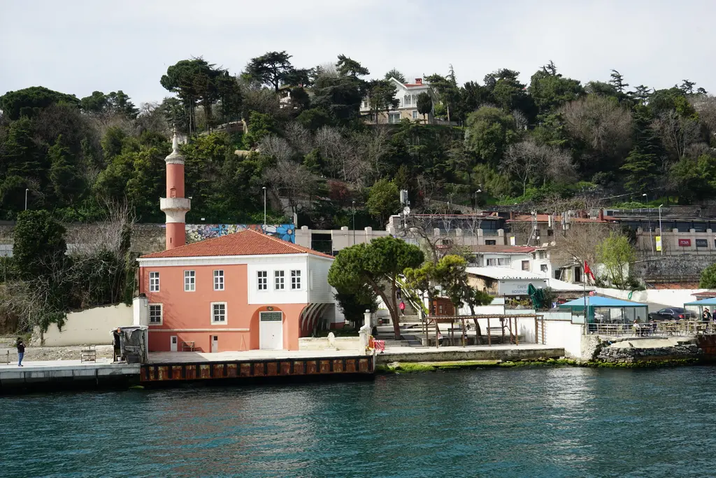 Boğaz'dan Defterdar İbrahim Paşa Camii; pembe cepheli küçük cami, ince minare, kıyı rıhtımı ve arkada ağaçlık yamaç.