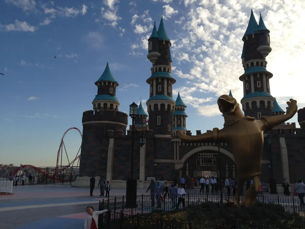 Vialand'de gün batımına doğru karşı ışıkta şato kuleleri; önde altın maskot heykeli, meydanda dolaşan kalabalık ve arkada roller coaster rayları.