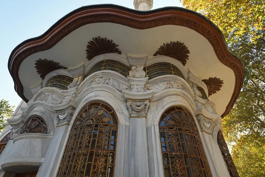 Nusretiye Camii'nin sebili; dalgalı saçaklı yuvarlak mermer yapı, yaldızlı parmaklıklı pencereler ve arkada sonbahar yaprakları.