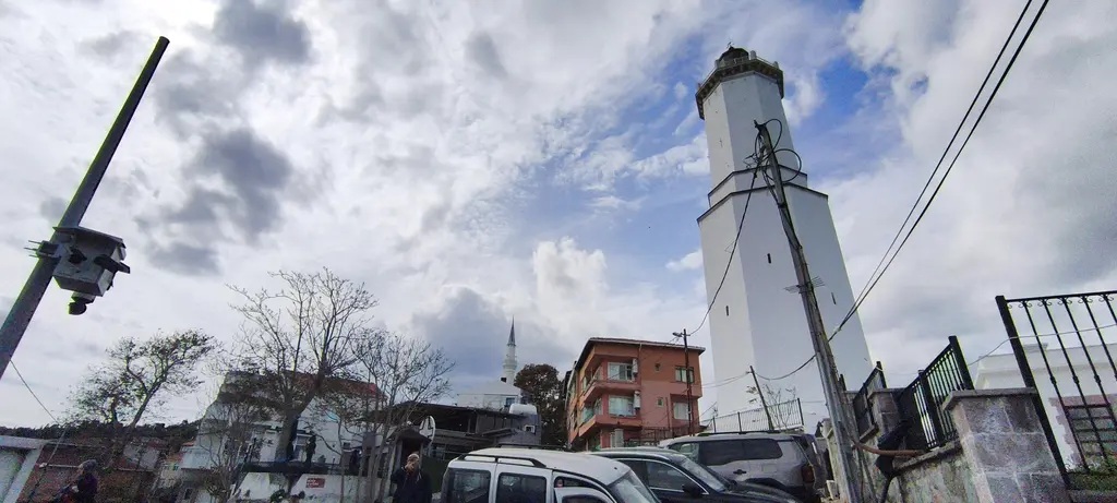 Rumelifeneri'nde fener; beyaz kule, demir çit, Türk bayrağı ve önde elektrik direğiyle sokak.