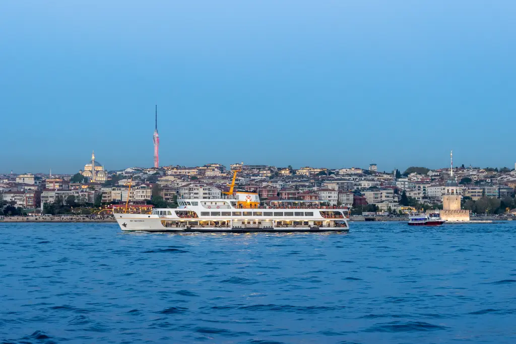 Denizden Üsküdar ve Kız Kulesi; önde beyaz vapur, kıyıda küçük kule, arkada yamaçlara yayılan evler ve camiler.