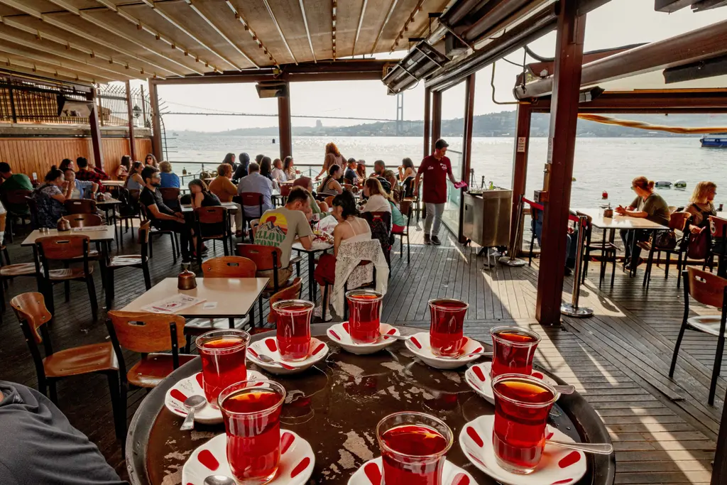Çınaraltı çay bahçesinde ince belli bardaklarla çay; dolu masalar, Boğaz manzarası ve uzakta köprü.