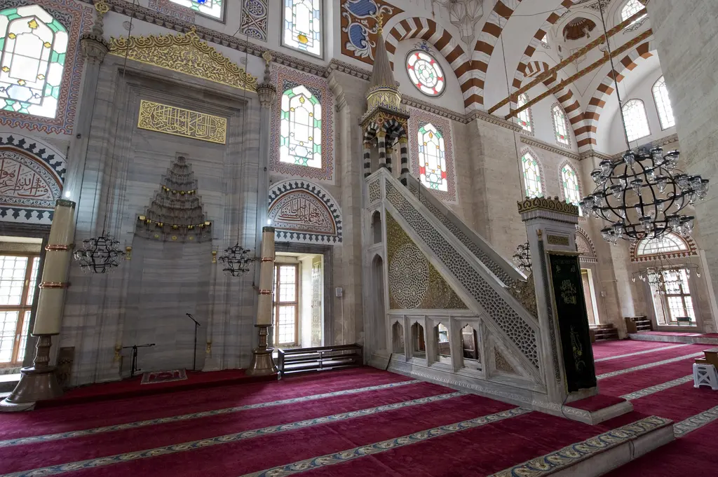 Şehzade Camii'nin kıble duvarı; mukarnas kavsaralı mermer mihrap, sağında mermer minber, iki yanda renkli cam pencereler ve önde koyu pembe halı.