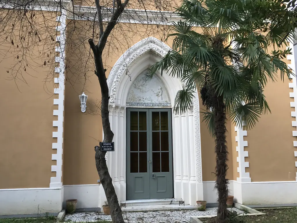 Surp Andon Ermeni Katolik Kilisesi; sarı cephe, beyaz sivri kemerli kapı, yanında palmiye ve ağaç.