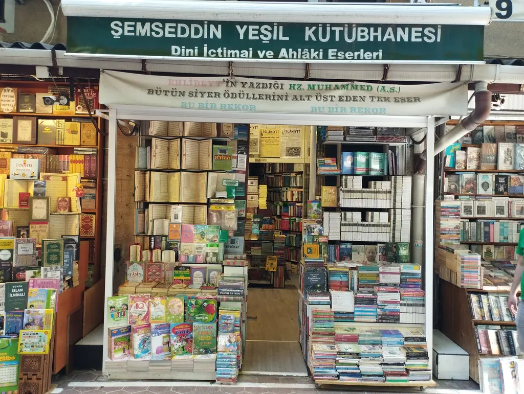 Sahaflar Çarşısı'nda koyu yeşil tabelalı bir kütüphane dükkânı; iki yanı ahşap vitrinli dar dükkân ağzı, açıkta sergilenen ciltli eserler ve önde renkli kitap standları.