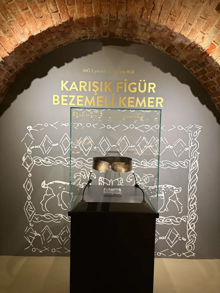 Karışık Figür Bezemeli Kemer; siyah kaide üstünde cam vitrin, arkada beyaz çizgiyle çizilmiş hayvan ve bitki motifleri ve tuğla kemer.