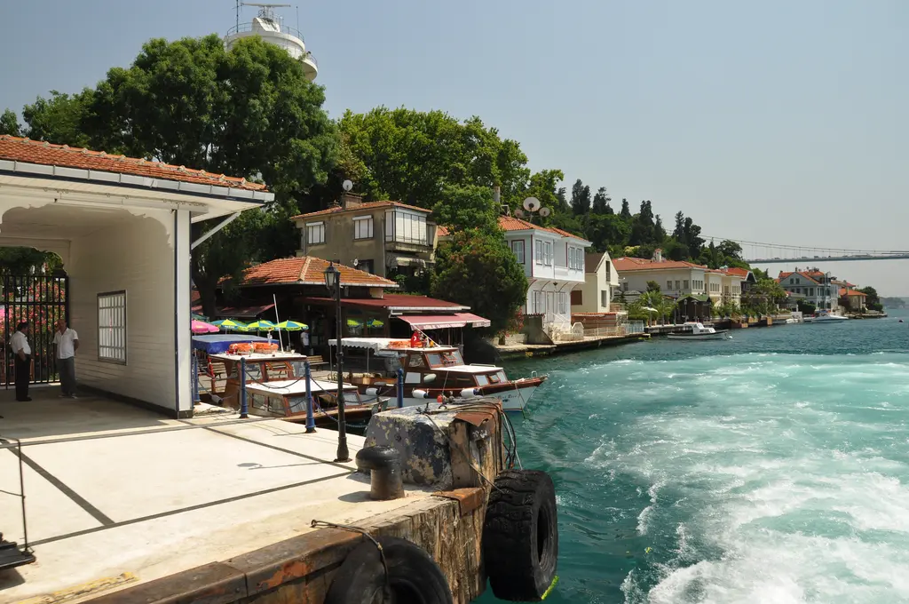 Kanlıca iskelesi; vapurdan görünen beyaz iskele binası, kıyıdaki şemsiyeli kafeler ve yamaçta evler.