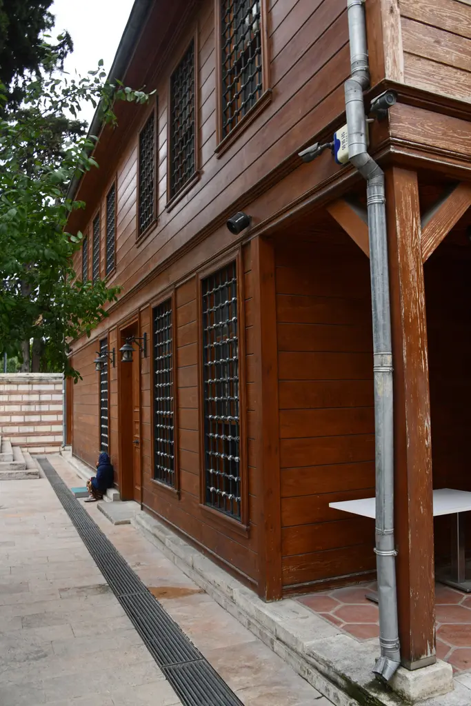 Şah Sultan Camii'nde ahşap bina; ahşap direkli giriş, demir şebekeli pencereler, yağmur borusu ve taş döşeli avlu.