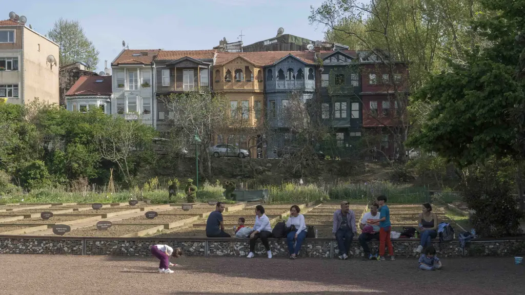 Kuzguncuk Bostanı'nda tarhların kenarında oturan insanlar; arkada renkli ahşap evler ve ağaçlar.
