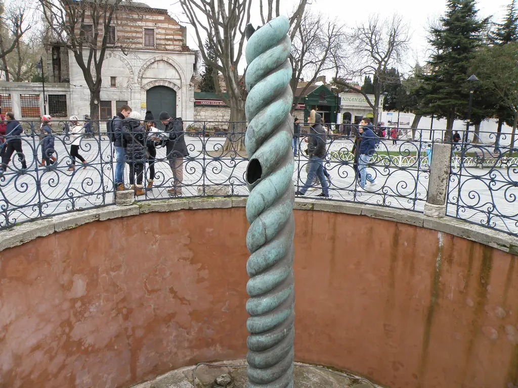 Yılanlı Sütun ve çevresindeki parmaklıklı çukur; pembe sıvalı iç duvar, arkada kubbeli taş bir yapı, çıplak ağaçlar ve meydanda yürüyenler.