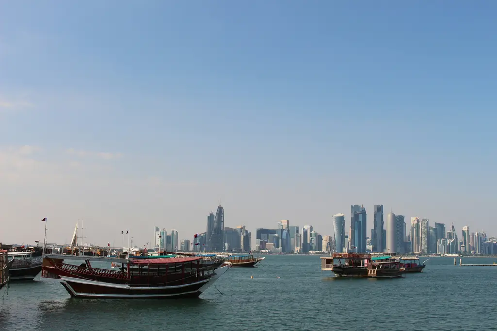 Doha Korniş'te ahşap dhow tekneleri; sıra hâlinde demirlemiş koyu renkli gövdeleri, arkada körfezin karşı yakasındaki gökdelenler ve açık gökyüzü.