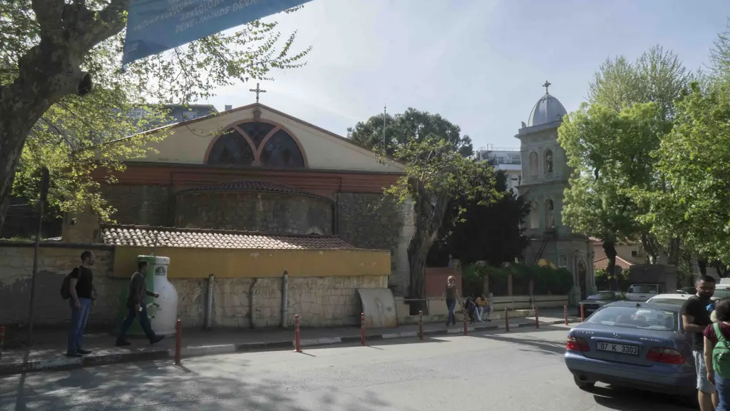 Kuzguncuk'ta Aya Pantelimon Kilisesi'nin çan kulesi; beyaz katlı kule, önde ağaçlar, kırmızı kapı ve yürüyen insanlar.