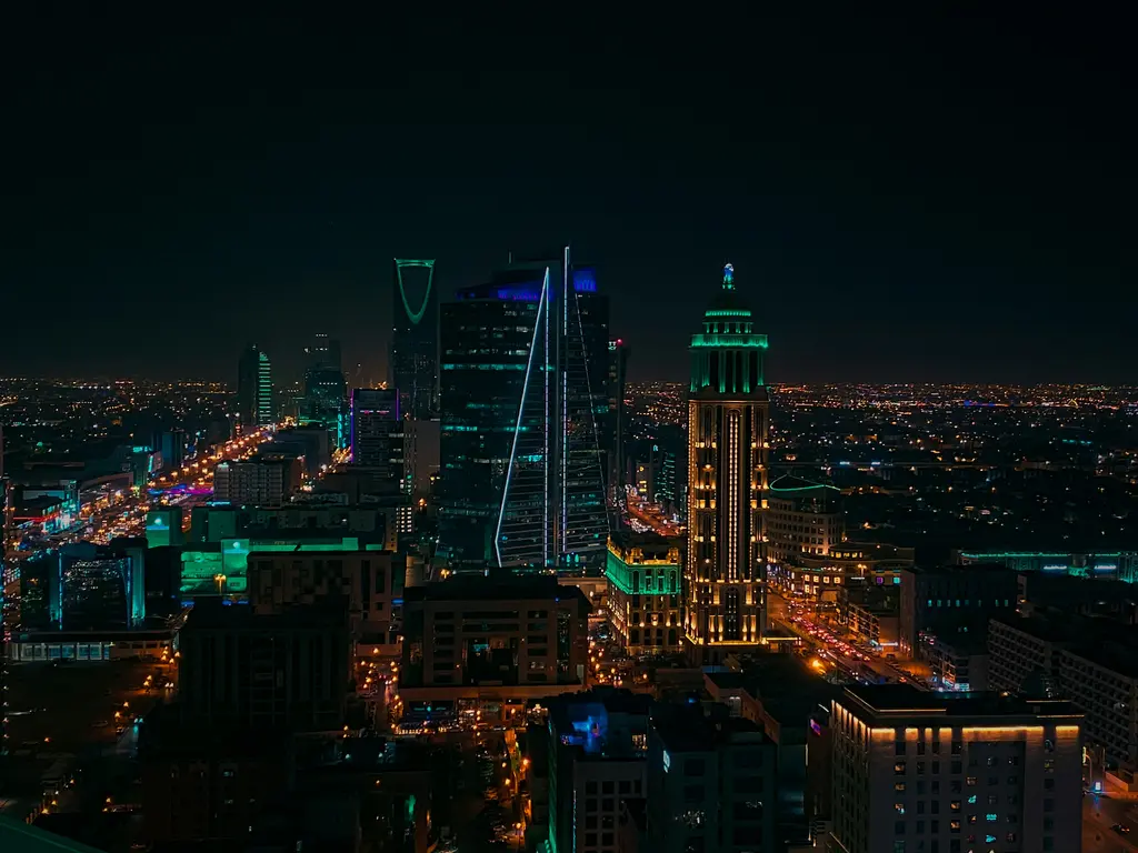 Riyad'da gece yukarıdan görünen şehir; yeşil ve mavi ışıkla aydınlatılmış gökdelenler, aralarındaki alçak binalar ve ufka uzanan cadde ışıkları.
