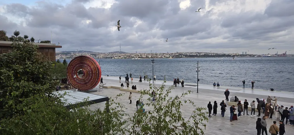 Galataport'ta kıyı meydanı; solda halkalardan oluşan büyük bakır renkli heykel, ortada kalabalık, arkada Boğaz ve martılar.