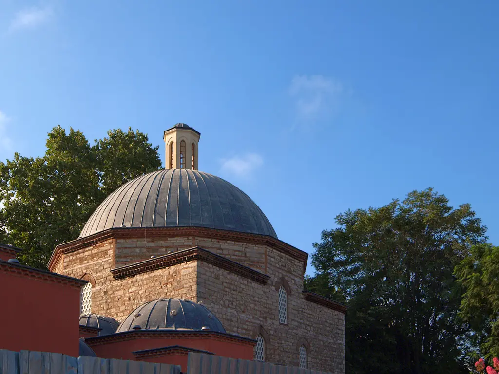 Mavi gökyüzüne karşı hamamın kurşun kaplı kubbesi; altında pembe sıvalı duvar, kenarda ikinci bir kubbe ve ağaç dalları.