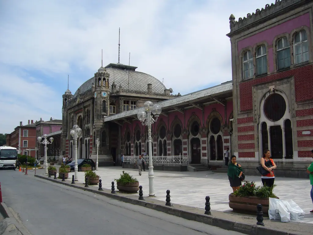 Sirkeci Garı'nın önündeki yaya yolu; sağda pembe duvar boyunca gül pencereler ve saçak, solda siyah babalar ile süslü beyaz lamba direkleri, ileride kubbeli saat kulesi bloğu.