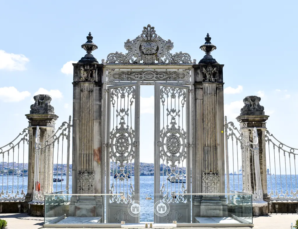 Dolmabahçe Sarayı'nda deniz kapısı ve parmaklıklar; dökme demir kanatlar, süslü taş direkler ve arkada Boğaz'ın suyu.