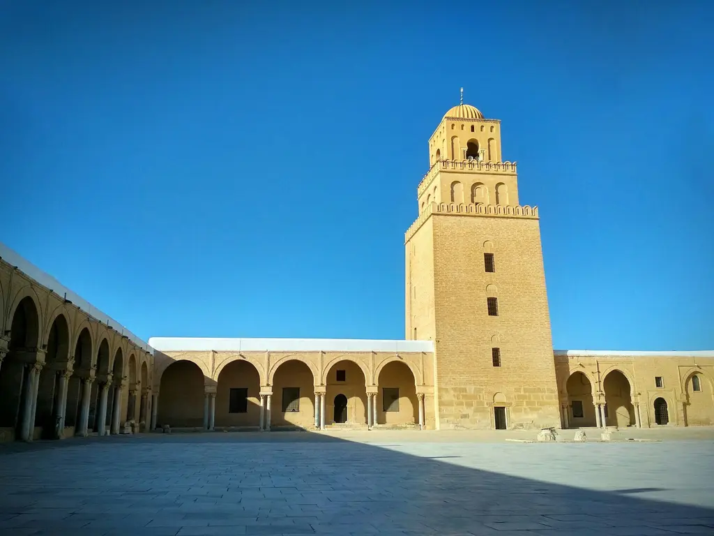 Tunus'ta Kairouan Ulu Camii; geniş taş avlu, kemerli revaklar ve arkada anıtsal kare minare, berrak mavi gökyüzü.