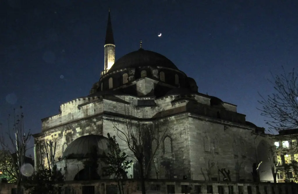 Gece Gazi Atik Ali Paşa Camii; aydınlatılmış taş cephe, karanlık kubbe, ışıklı minare, gökyüzünde hilal ve önde ağaçlar.