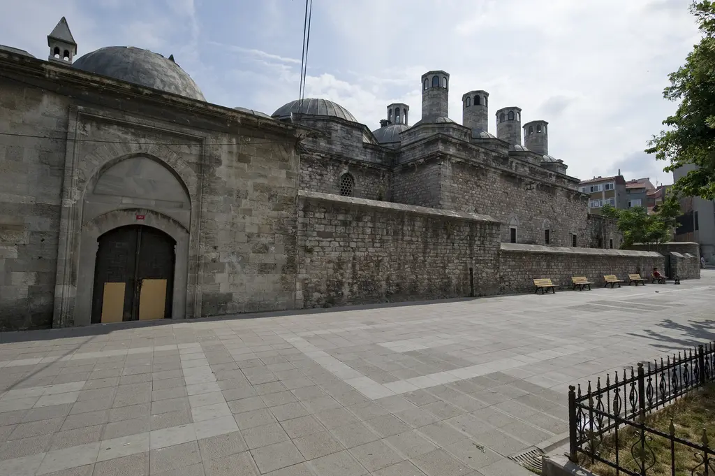 Haseki Sultan Külliyesi; sivri kemerli ahşap kapı, uzun kesme taş duvar, arkada bacalar ve kubbeler, önde taş döşeli geniş yol.