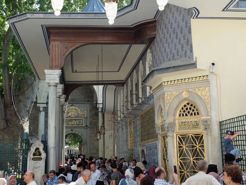 Eyüp Sultan Türbesi'nin girişi; saçaklı revak, yaldızlı sebil cephesi, mermer sütunlar ve yoğun ziyaretçi kalabalığı.