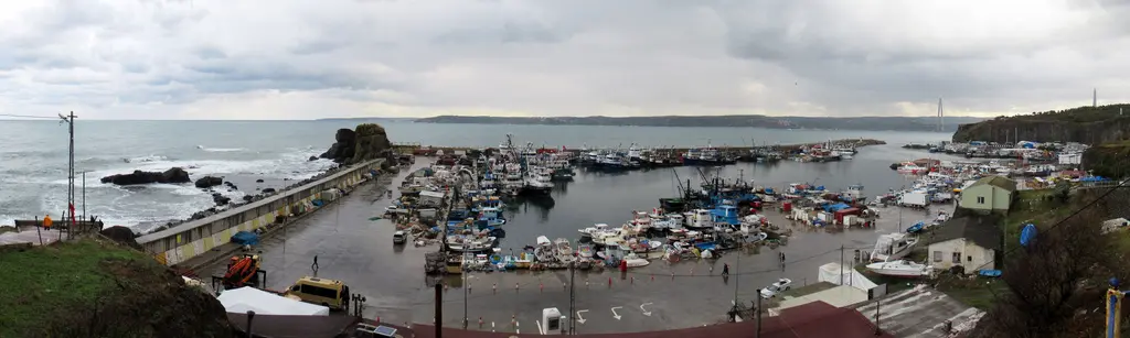 Rumelifeneri balıkçı limanı; dalgakıranın içinde sıra sıra tekneler, kıyıda kayalar ve arkada deniz.