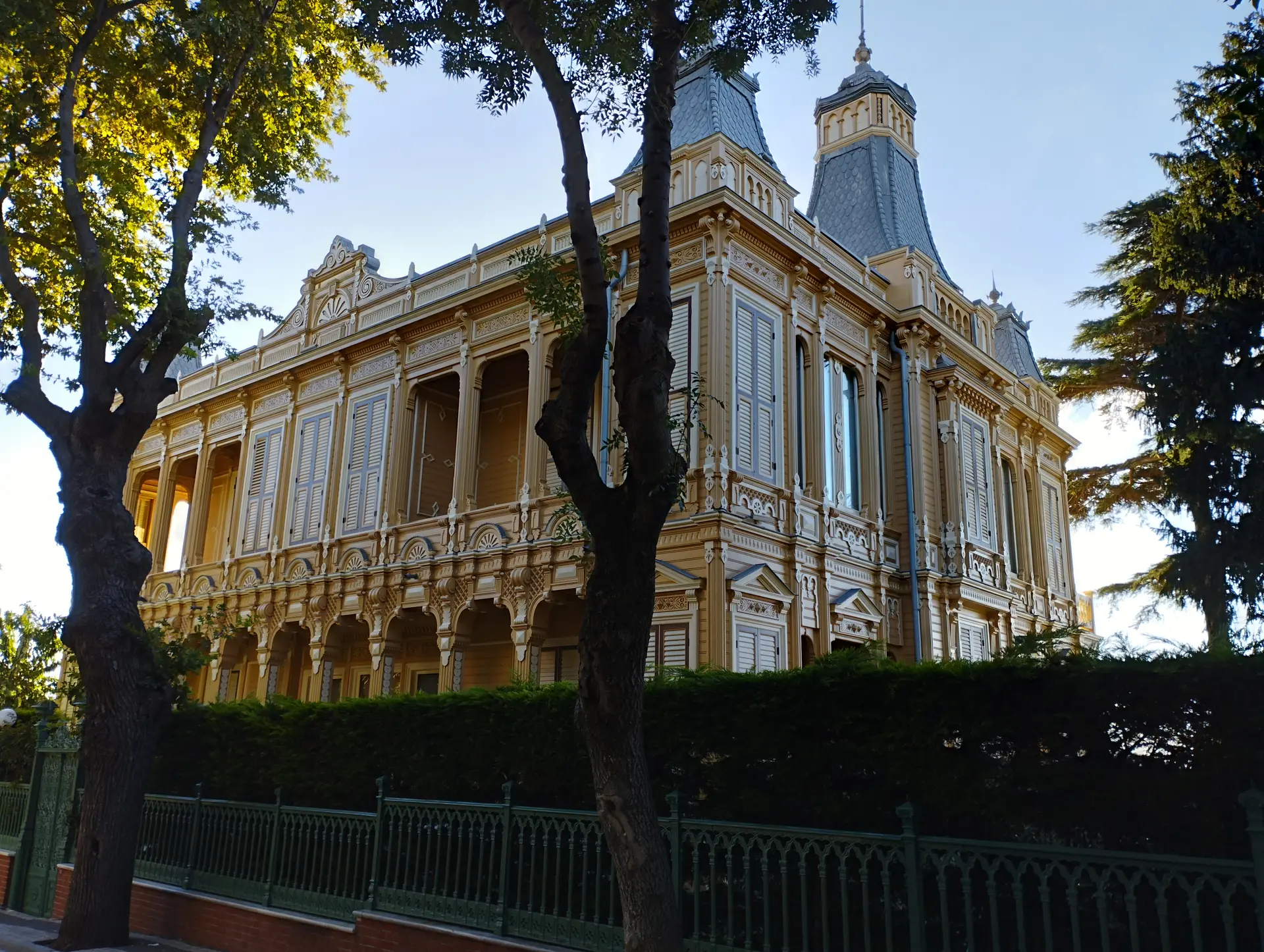 Büyükada'da köşk; sarı cepheli, kuleli büyük konak, demir parmaklıklı bahçe ve önünde yaşlı ağaçlar.