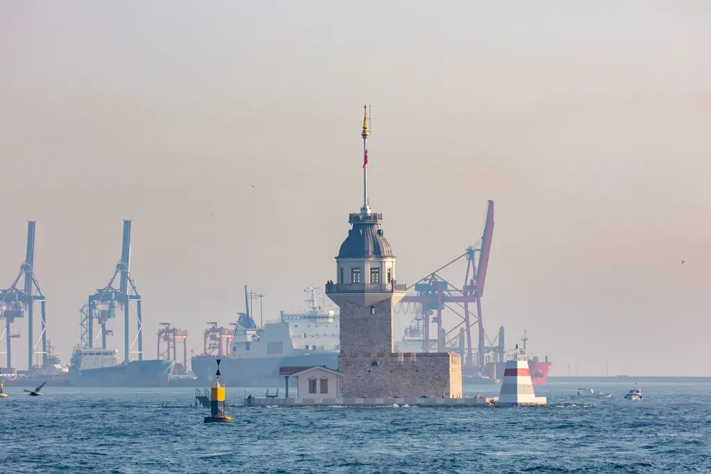 Kız Kulesi; puslu havada taş kule, arkasında Haydarpaşa limanının vinçleri ve önde dalgalı deniz.