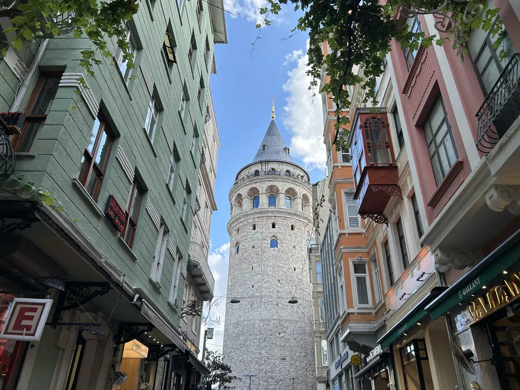 Galata'da dar bir sokağın ucunda Galata Kulesi; iki yanda renkli cepheli eski evler, sarkan yapraklar ve dükkân tabelaları.