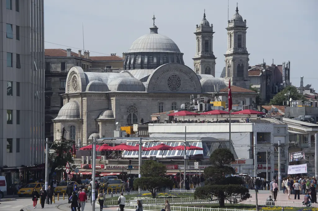 Taksim Meydanı'ndan Aya Triada Kilisesi; önde dükkânlar ve kalabalık, arkada kubbe ile iki çan kulesi, yanında apartmanlar.