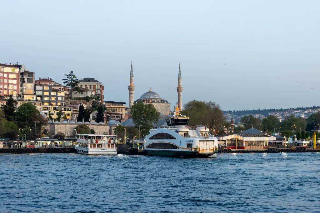 Denizden Üsküdar; kıyıda iskele yapıları ve vapurlar, arkada çift minareli cami ve yamaca yayılan binalar.