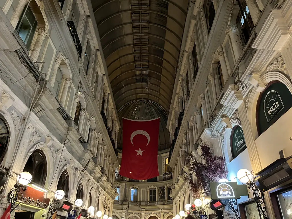 Çiçek Pasajı'nda geçit; tonozdan sarkan büyük Türk bayrağı, iki yanda süslü beyaz cepheler, yuvarlak lambalar ve meyhane tabelaları.