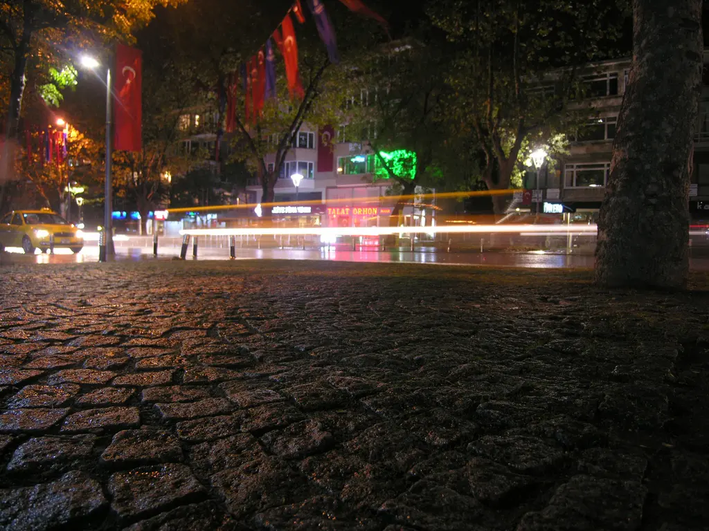 Gece Bağdat Caddesi'nde taş döşeli kaldırım; geçen arabaların ışık izleri, çınarlar ve bayraklar.
