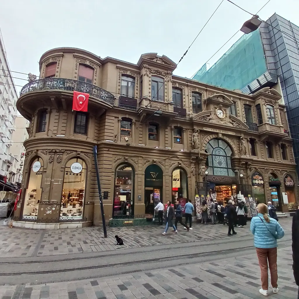 İstiklal Caddesi'nde Çiçek Pasajı'nın köşesi; yarım yuvarlak köşe kulesi, balkonda Türk bayrağı, zemin katta dükkânlar ve önden geçen yayalar.