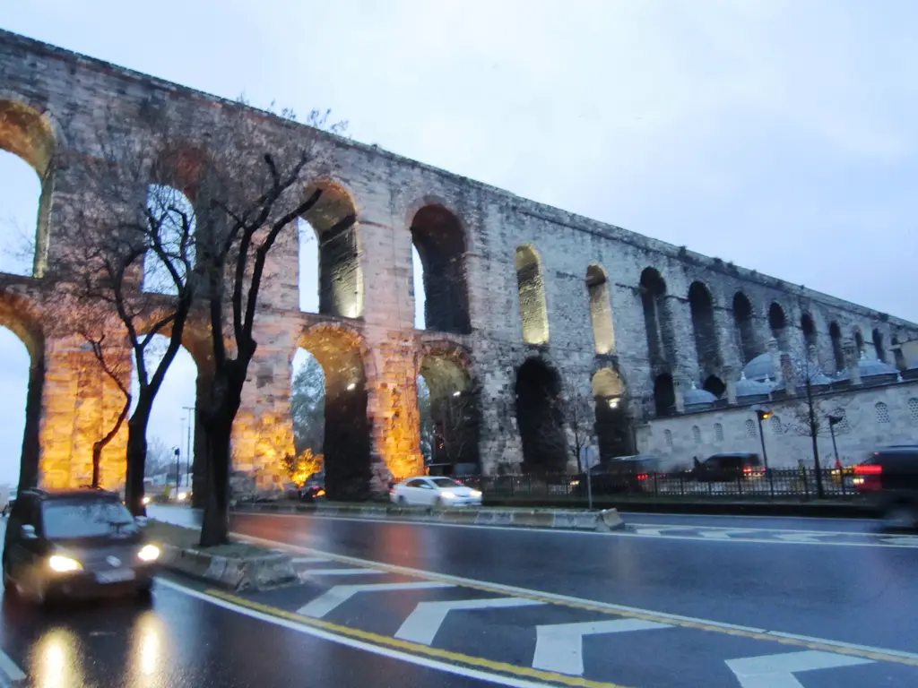 Bozdoğan Kemeri alacakaranlıkta, yağmurdan sonra; alt kemerler turuncu projektörle aydınlanmış, üst kısım hâlâ günün mavisinde, ıslak asfaltta sarı yol çizgileri parlıyor.