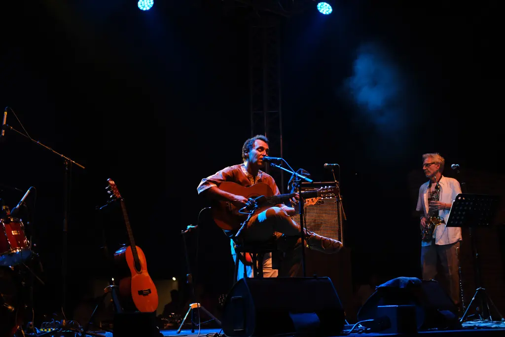 Esma Sultan Yalısı'ndaki sahnede müzisyenler; gitar çalan şarkıcı, saksafoncu, mikrofonlar ve mavi ışıklar.